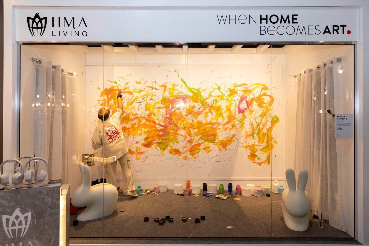 HMA Living gallery window under a When Home Becomes Art fascia, an artist painting a mural live on the back wall