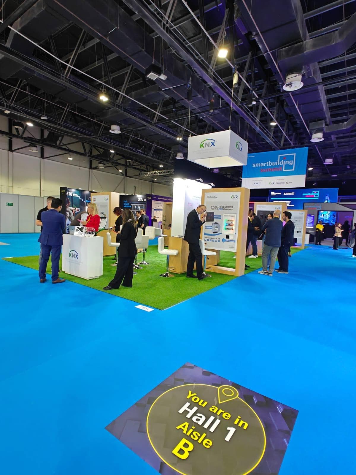 KNX stand at the Smart Building Summit, a suspended logo cube over an artificial-lawn floor and demo panels