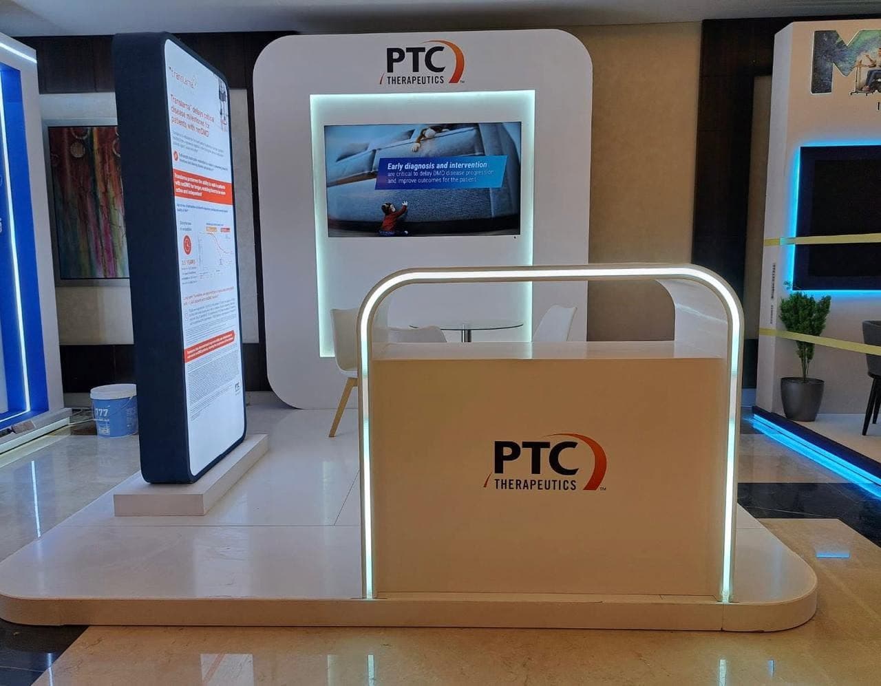 PTC Therapeutics stand, a backlit screen recess and a curved light-line counter on a stone-tiled floor